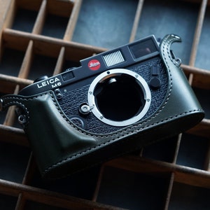 Könnte beinhalten: Schwarze Leica M6 Kamera mit grüner Lederhülle. Die Kamera hat einen silbernen Objektivanschluss und ein rotes Leica-Logo. Sie liegt auf einem Holztablett mit mehreren Fächern.