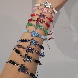 以下が含まれることがあります： カラフルなビーズとタートルチャームが付いたブレスレットのセット。各ブレスレットは、異なる色の紐とビーズでできており、カラフルなデザインのシルバータートルチャームが付いています。