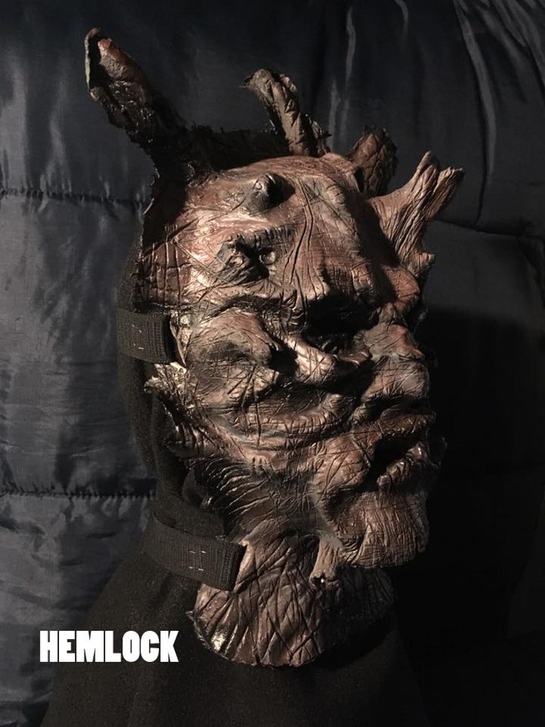 May include: A detailed, brown Hemlock mask with a textured, wood-like appearance. The mask features prominent, branch-like protrusions and is secured with a black strap. The word "HEMLOCK" is displayed in white text at the bottom.