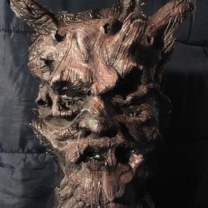 May include: A detailed, textured mask with a dark brown and reddish-brown color scheme. The mask has a gnarled, tree-like appearance with prominent horns and the word "HEMLOCK" in white text at the bottom.