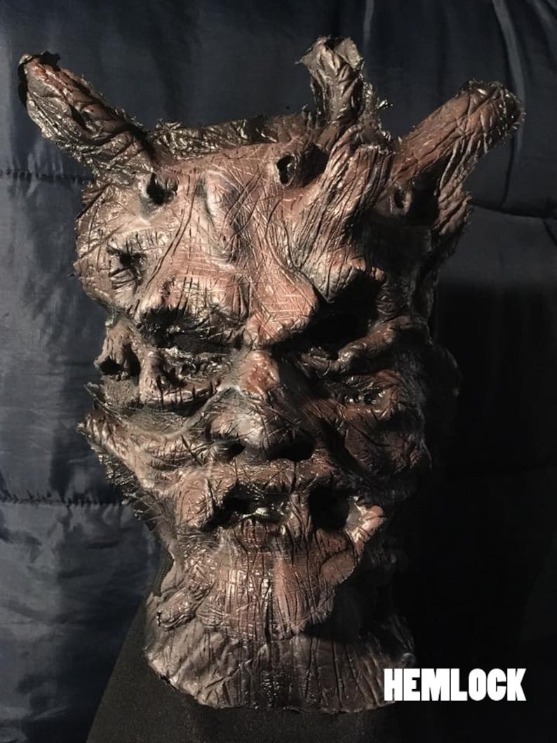 May include: A detailed, textured mask with a dark brown and reddish-brown color scheme. The mask has a gnarled, tree-like appearance with prominent horns and the word "HEMLOCK" in white text at the bottom.