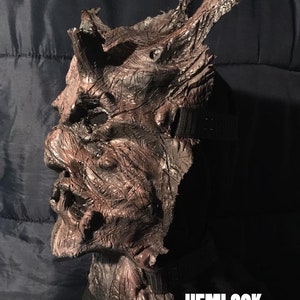 May include: A brown and black Hemlock mask with a textured, weathered appearance. The mask has a detailed, gnarled design with horns and a strap for wearing. The word "HEMLOCK" is printed in white.
