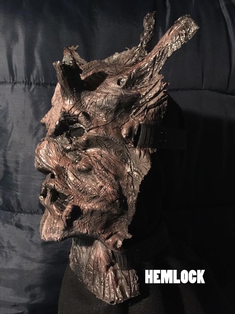 May include: A brown and black Hemlock mask with a textured, weathered appearance. The mask has a detailed, gnarled design with horns and a strap for wearing. The word "HEMLOCK" is printed in white.