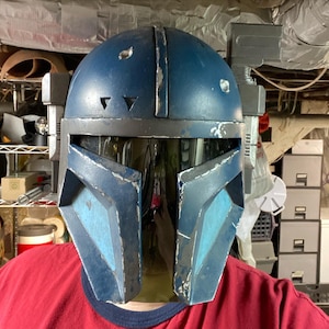 Pode incluir: Um capacete Mandaloriano azul e cinza desgastado com uma viseira fumê. O capacete apresenta elementos de design angulares e desgaste visível, sugerindo uma estética desgastada pela batalha. O capacete tem um acessório retangular na lateral.