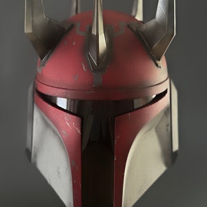 Puede incluir: Un casco rojo y blanco con una visera negra y tres cuernos negros en la parte superior. El casco tiene un aspecto desgastado y descolorido.