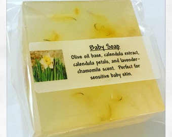 May include: A clear, rectangular bar of baby soap, packaged in clear plastic. The soap is a pale yellow color and contains small, visible flower petals. A label reads "Baby Soap" and lists ingredients like olive oil, calendula, and chamomile.