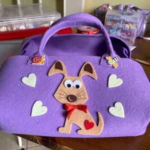 Può includere: Borsa a mano in feltro viola con un'applicazione di cane in feltro marrone, un cuore rosso e cuori in feltro bianco. La borsa ha un manico ed è decorata con una lumaca e una libellula.