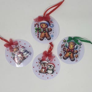 Può includere: Quattro addobbi natalizi rotondi con diversi disegni festivi. Gli addobbi sono bianchi con nastro rosso e verde. I disegni includono un uomo di pan di zenzero, una renna e un pinguino, una renna e un orso polare, e un uomo di pan di zenzero con cuffie.