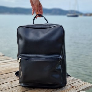 May include: A black leather backpack with a zippered front pocket. The backpack is sitting on a wooden surface.