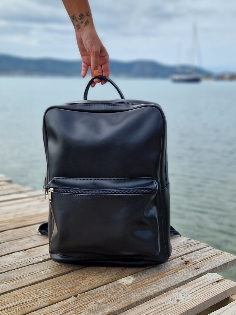 May include: A black leather backpack with a zippered front pocket. The backpack is sitting on a wooden surface.
