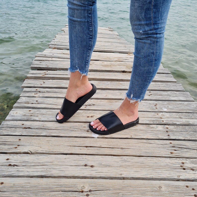 May include: A pair of black leather slides with a rubber sole. The slides are worn by a person standing on a wooden dock.