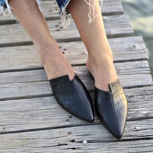 May include: A pair of black leather mules with a crocodile texture. The shoes have a pointed toe and a low heel. The image shows the shoes on a wooden surface.