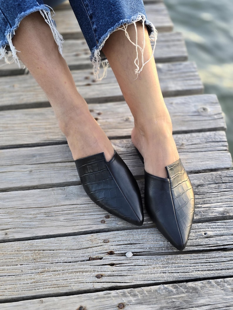 May include: A pair of black leather mules with a crocodile texture. The shoes have a pointed toe and a low heel. The image shows the shoes on a wooden surface.