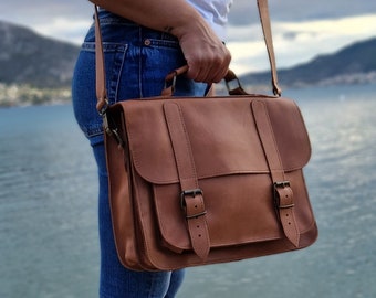 May include: A brown leather satchel bag with two buckles and a long adjustable strap. The bag is worn over the shoulder of a person wearing blue jeans.