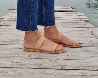 May include: Tan leather sandals with a simple design. The sandals have a strap around the ankle with a buckle and a strap across the toes. The sandals are worn with blue jeans. The background is a wooden dock.