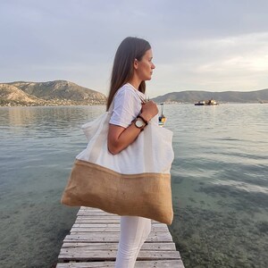 May include: A woman standing on a wooden dock by the water, holding a large white and brown tote bag. The bag has a white canvas top and a brown burlap bottom.