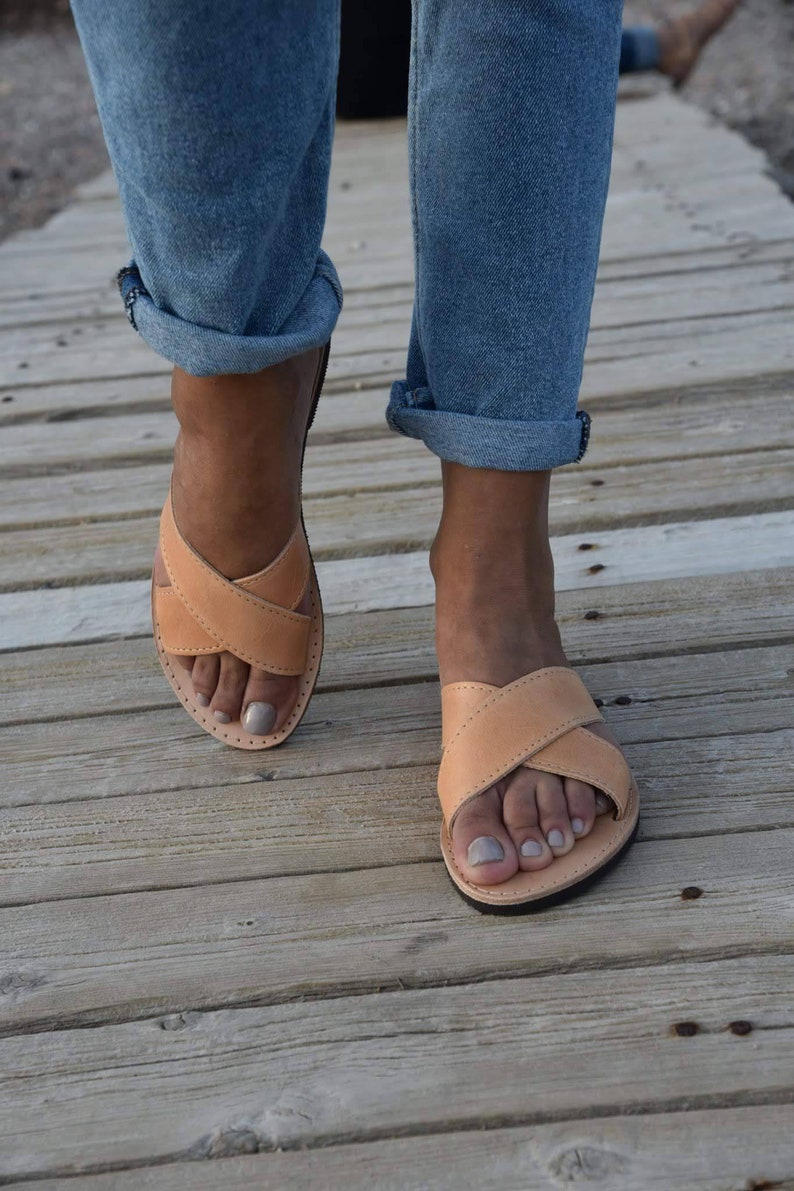 May include: A pair of tan leather sandals with criss-cross straps. The sandals are worn by a person wearing blue jeans.