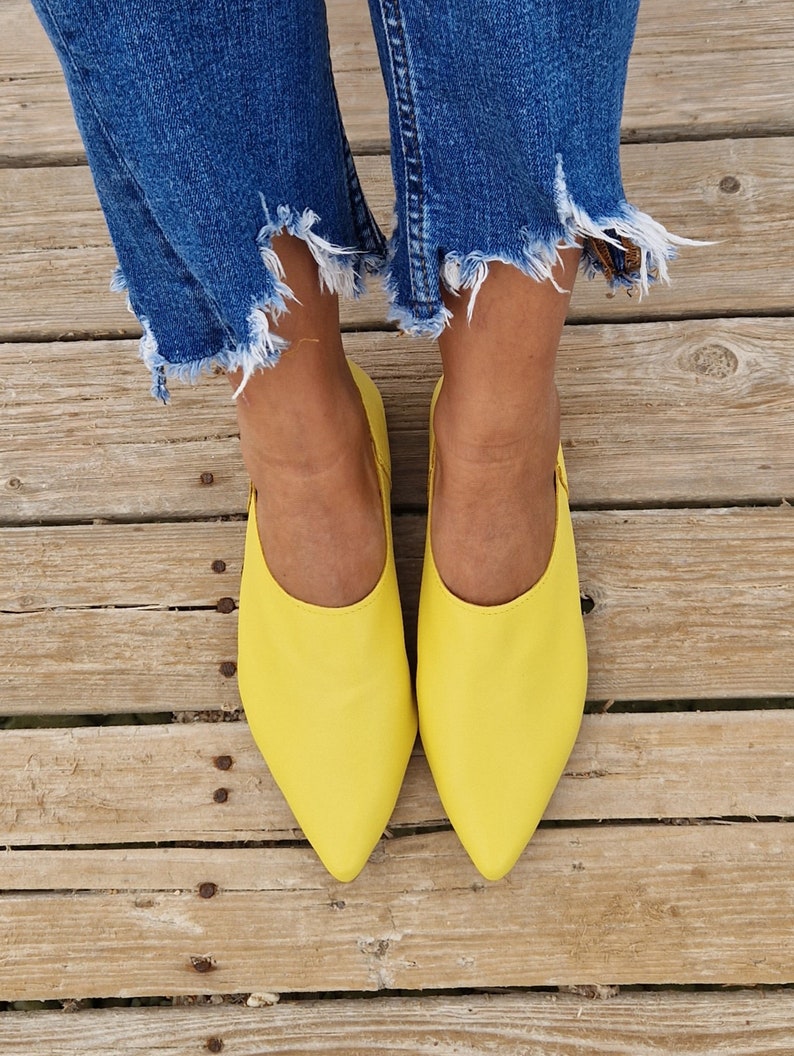 May include: A pair of bright yellow pointed-toe flats with a simple design. The shoes are worn by a person wearing blue jeans.