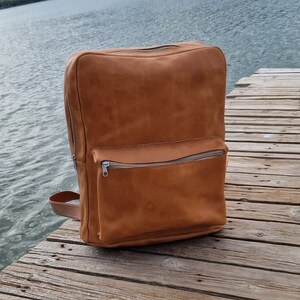 May include: A brown leather backpack with a zippered front pocket. The backpack is sitting on a wooden surface.