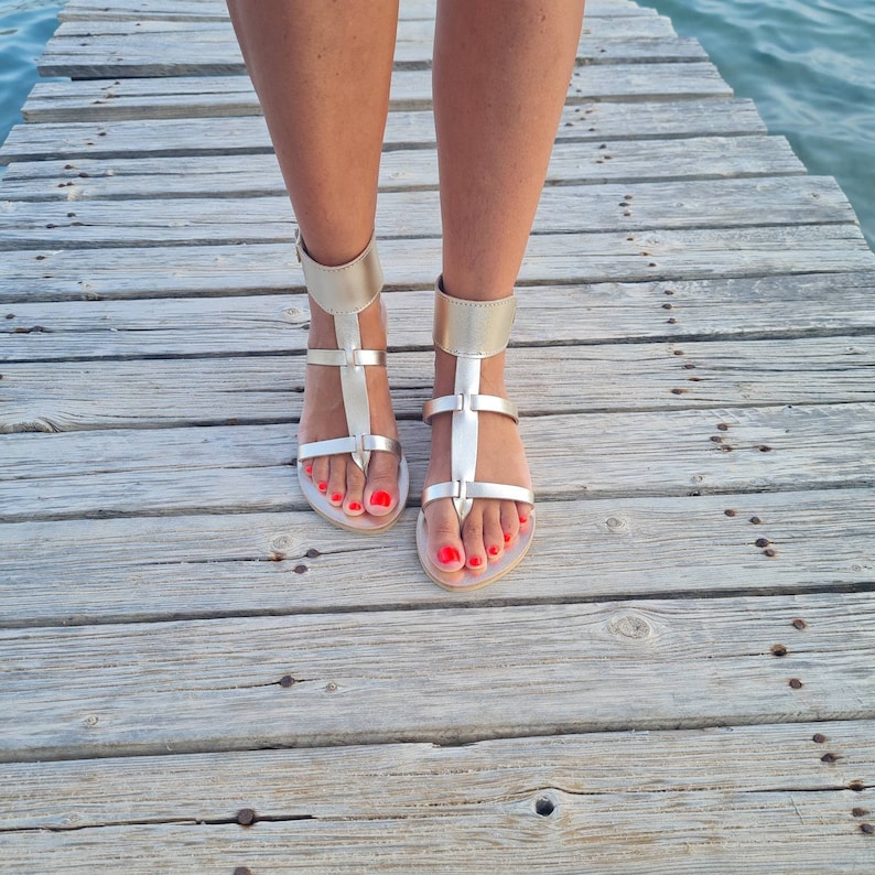 May include: A pair of gold leather sandals with ankle straps. The sandals have a simple design with a toe strap and a buckle closure at the ankle. The sandals are worn on a wooden surface.