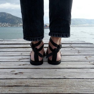K&ouml;nnte beinhalten: Schwarze Ledersandalen mit mehreren Riemen und einem Schnallenverschluss. Die Sandalen werden von einer Person getragen, die auf einem Holzsteg steht.