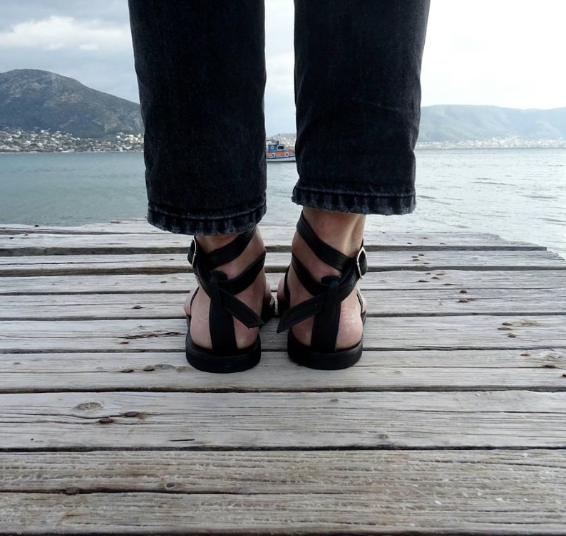 K&ouml;nnte beinhalten: Schwarze Ledersandalen mit mehreren Riemen und einem Schnallenverschluss. Die Sandalen werden von einer Person getragen, die auf einem Holzsteg steht.