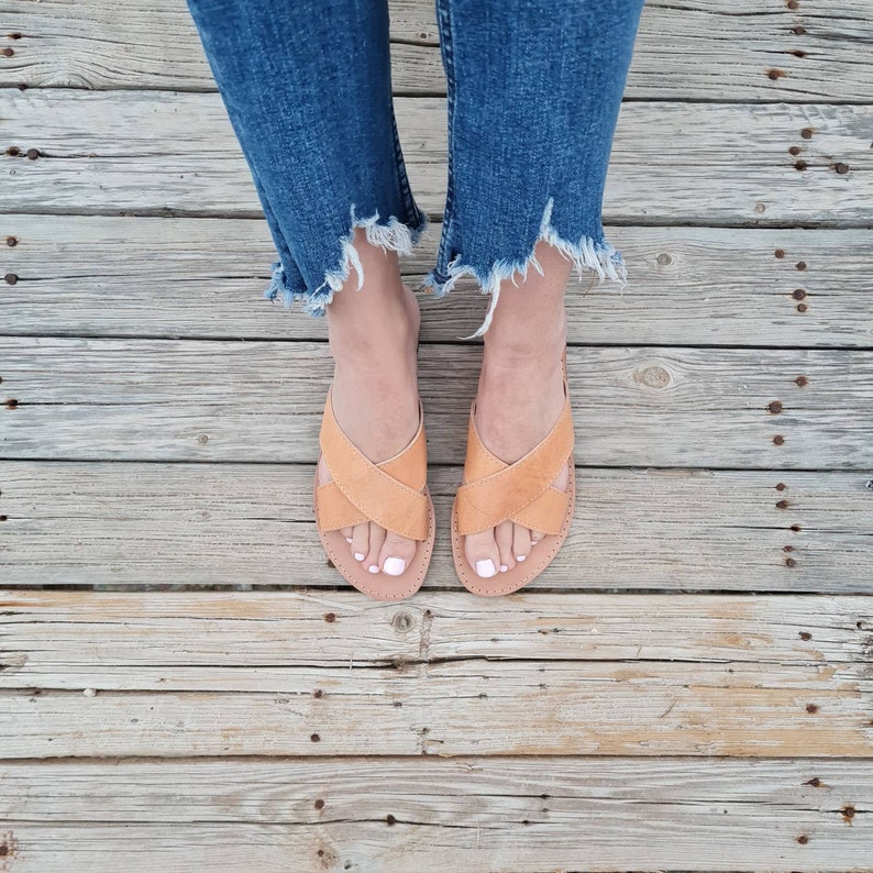 May include: A pair of tan leather sandals with criss-cross straps. The sandals are worn by a person wearing blue jeans.