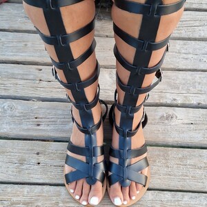 Op de afbeelding: Zwarte gladiatorsandalen van leer met meerdere banden die tot aan het been reiken. De sandalen hebben een platte zool en worden gedragen op een houten oppervlak.