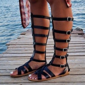 Op de afbeelding: Zwarte gladiatorsandalen van leer met meerdere banden en gespen. De sandalen hebben een platte zool en worden gedragen op een houten pier.