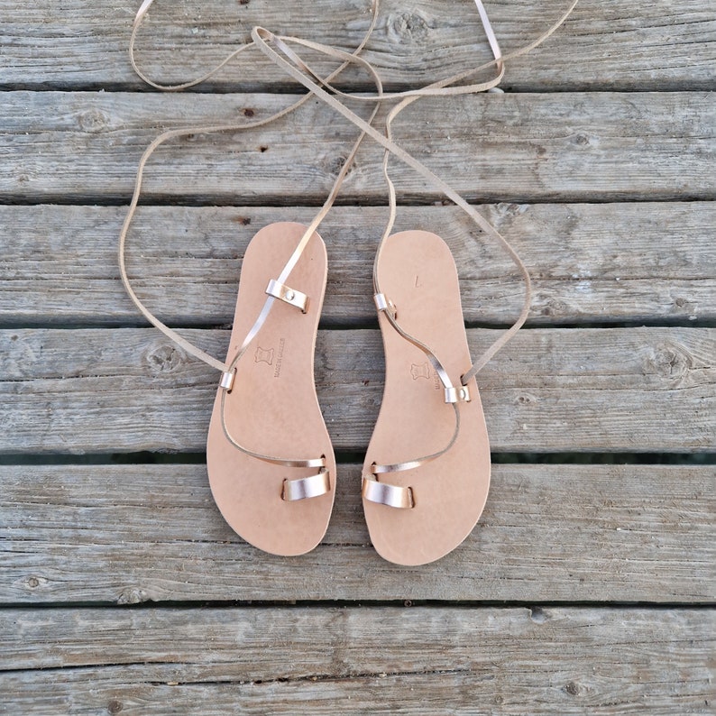 May include: A pair of light brown leather sandals with rose gold metallic straps. The sandals have a toe loop and ankle straps that wrap around the leg. The sandals are displayed on a weathered wooden surface.