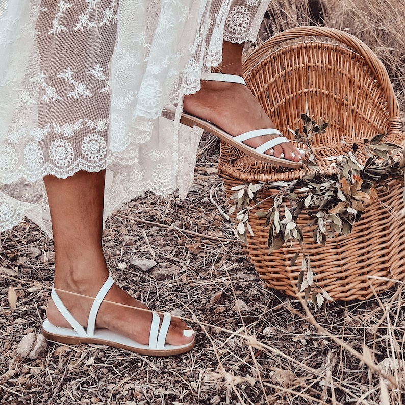 May include: A pair of white leather sandals with thin straps. The sandals are worn by a person standing on a wicker basket.