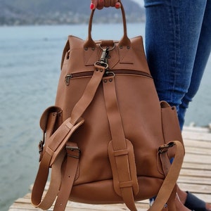 Puede incluir: Una mochila de cuero marr&oacute;n con correas ajustables y varios bolsillos. La mochila tiene un asa superior y un cierre de cremallera.