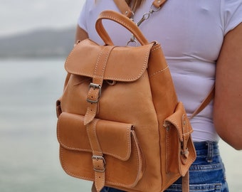 May include: A tan leather backpack with a flap closure and a front pocket. The backpack has adjustable straps and a buckle closure.