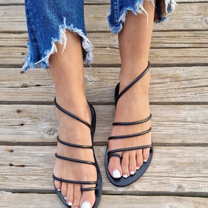 May include: Black leather sandals with multiple straps wrapping around the foot and ankle. The sandals have a flat sole and are worn on a wooden surface.
