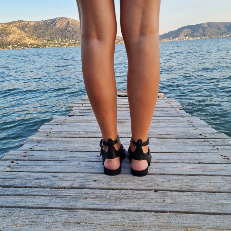 May include: A pair of black leather sandals with ankle straps. The sandals are worn by a person standing on a wooden dock overlooking a body of water.