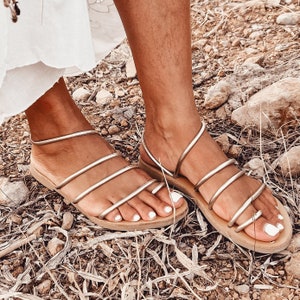 Peut inclure: Une paire de sandales en cuir dorées à plusieurs lanières. Les sandales sont portées aux pieds d'une personne et sont posées sur un fond d'herbe sèche et de rochers.