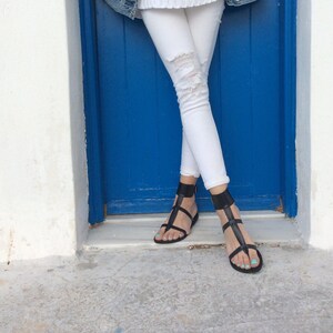 May include: A pair of black leather sandals with ankle straps. The sandals are worn by a person standing in front of a blue door.