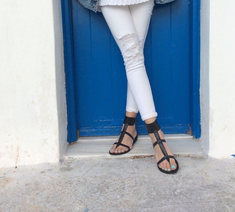 May include: A pair of black leather sandals with ankle straps. The sandals are worn by a person standing in front of a blue door.