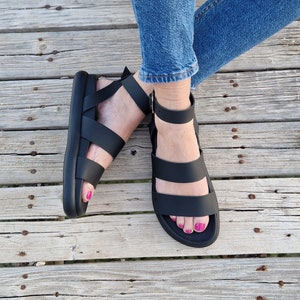 May include: Black leather sandals with two straps and an ankle strap. The sandals have a thick platform sole.