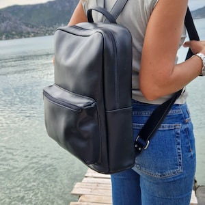 May include: A black leather backpack with a zippered front pocket. The backpack is worn by a person standing on a wooden dock.