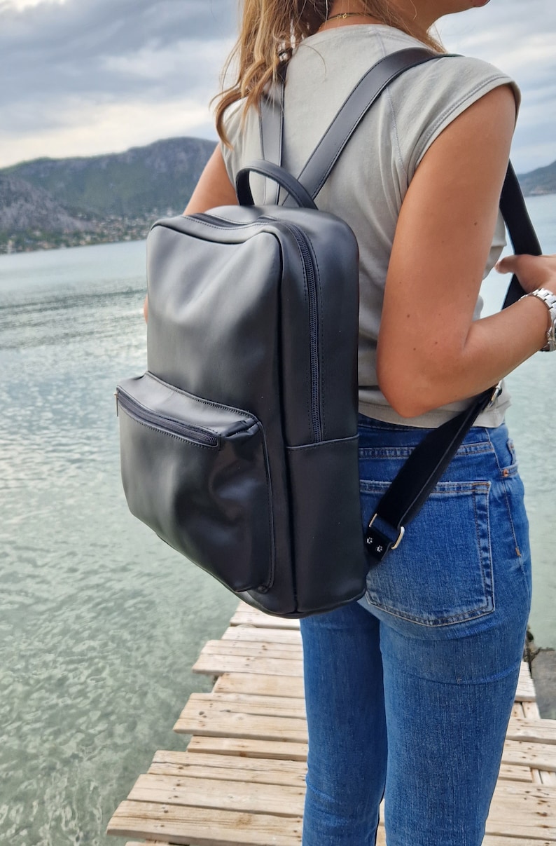 May include: A black leather backpack with a zippered front pocket. The backpack is worn by a person standing on a wooden dock.