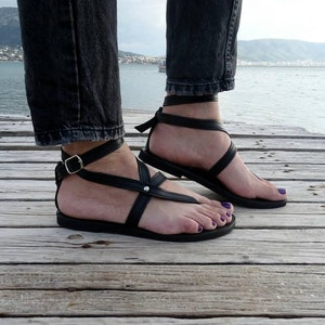 K&ouml;nnte beinhalten: Schwarze Ledersandalen mit Kn&ouml;chelriemen und einer Schnalle. Die Sandalen werden auf einem Holzsteg mit Wasser im Hintergrund getragen.