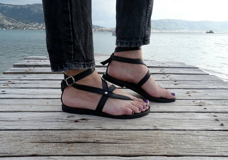 K&ouml;nnte beinhalten: Schwarze Ledersandalen mit Kn&ouml;chelriemen und einer Schnalle. Die Sandalen werden auf einem Holzsteg mit Wasser im Hintergrund getragen.