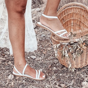 May include: A pair of white leather sandals with thin straps. The sandals are on a woman's feet and the woman is standing on a grassy surface. A wicker picnic basket is in the background.