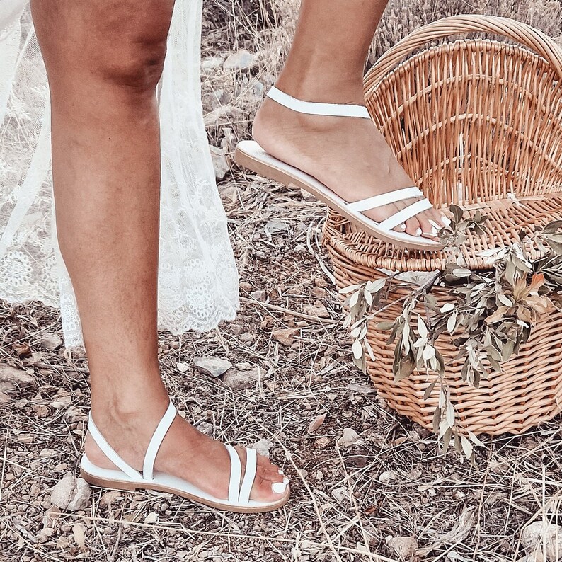 May include: A pair of white leather sandals with thin straps. The sandals are on a woman's feet and the woman is standing on a grassy surface. A wicker picnic basket is in the background.