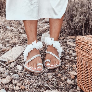 May include: White sandals with flower embellishments and pearl accents. The sandals have a toe strap and an ankle strap.