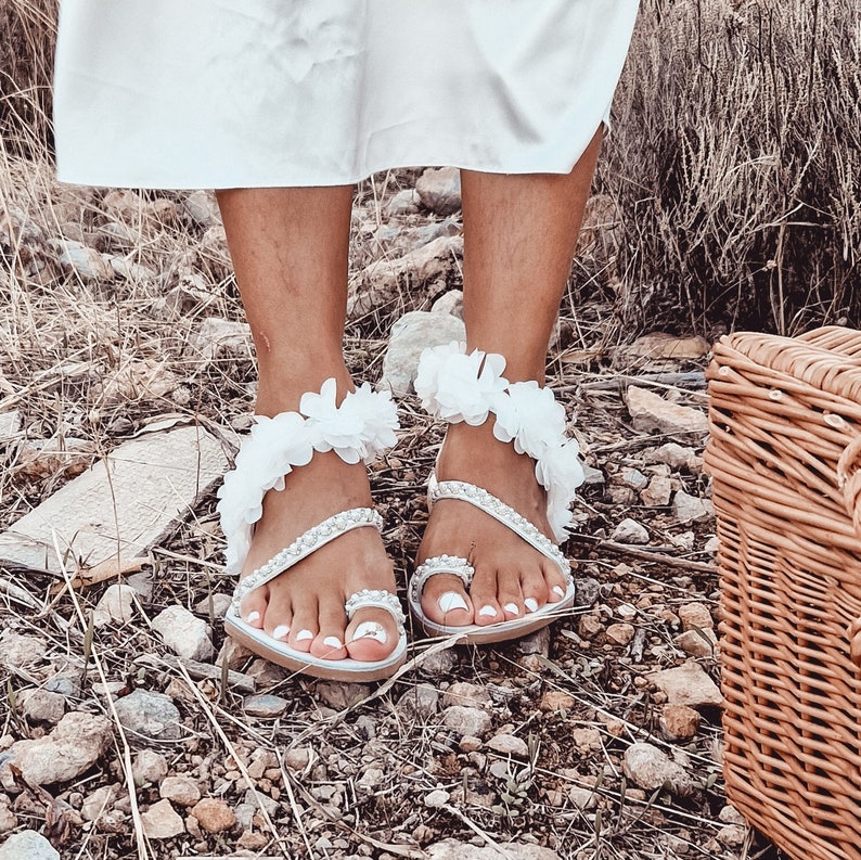May include: White sandals with flower embellishments and pearl accents. The sandals have a toe strap and an ankle strap.