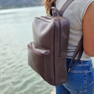 May include: A brown leather backpack with a zippered front pocket. The backpack is being worn by a person standing near a body of water.