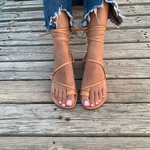 May include: Tan leather sandals with wrap-around ankle straps. The sandals feature a toe loop design and are worn with distressed denim jeans. The person's toenails are painted a light pink color. The sandals are on a wooden surface.