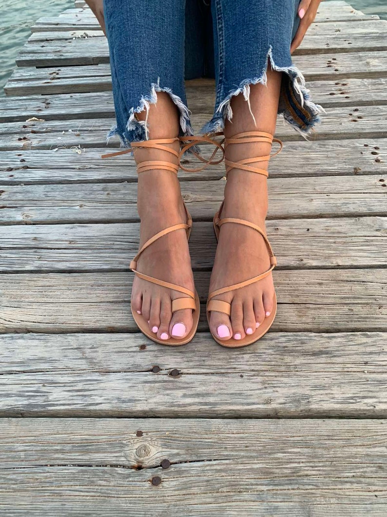 May include: Tan leather sandals with wrap-around ankle straps. The sandals feature a toe loop design and are worn with distressed denim jeans. The person's toenails are painted a light pink color. The sandals are on a wooden surface.